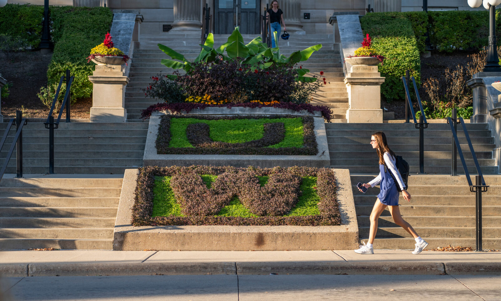 Students and pedestrians walk past contrasting green and brown plants form the letters U and W in flowerbeds